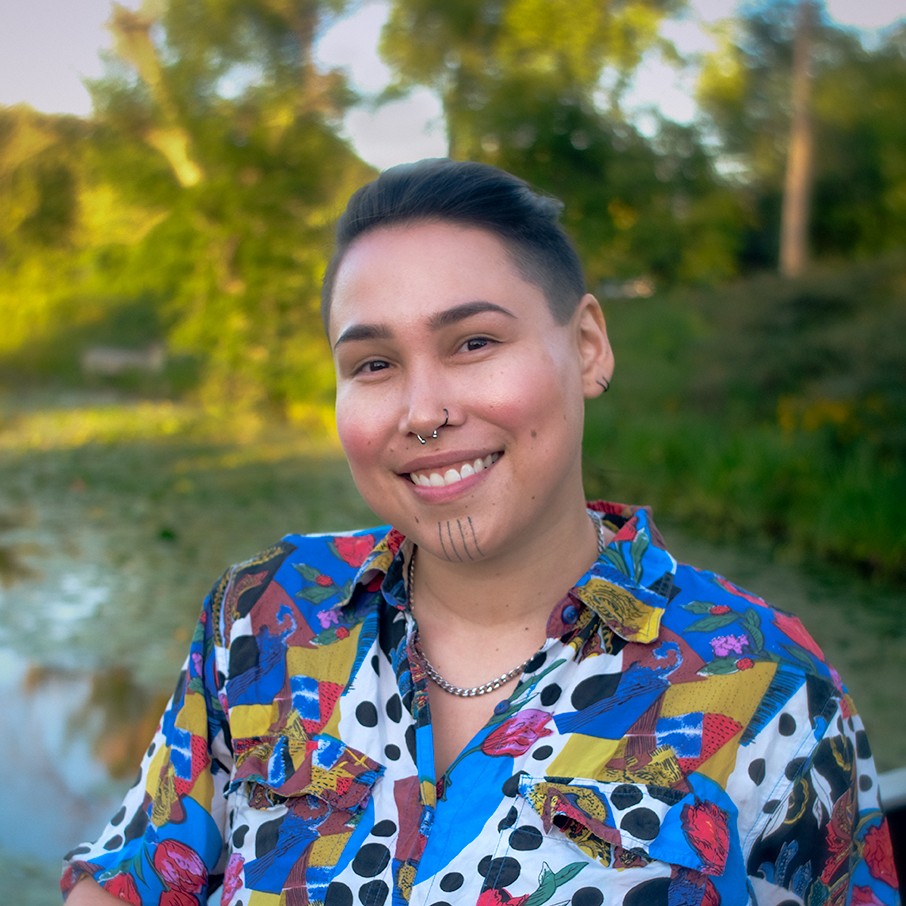 A square portrait of Nikita smiling in a vibrant button up shirt, with a stream and foliage in the background